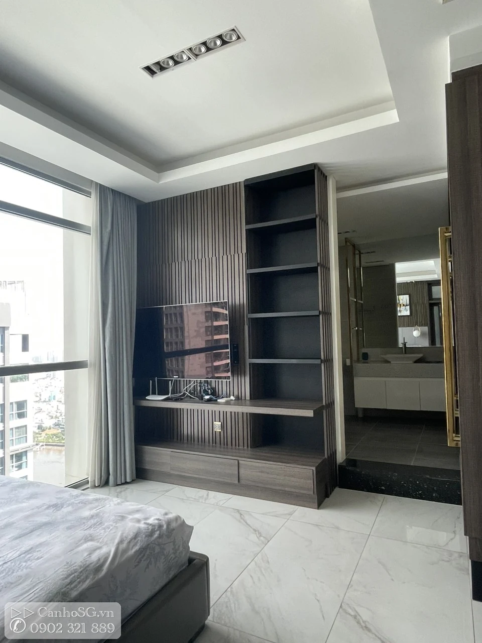 Phòng ngủ master rộng với kệ TV, tủ trưng bày và cửa mở vào khu lavabo, đón sáng tự nhiên từ cửa kính lớn