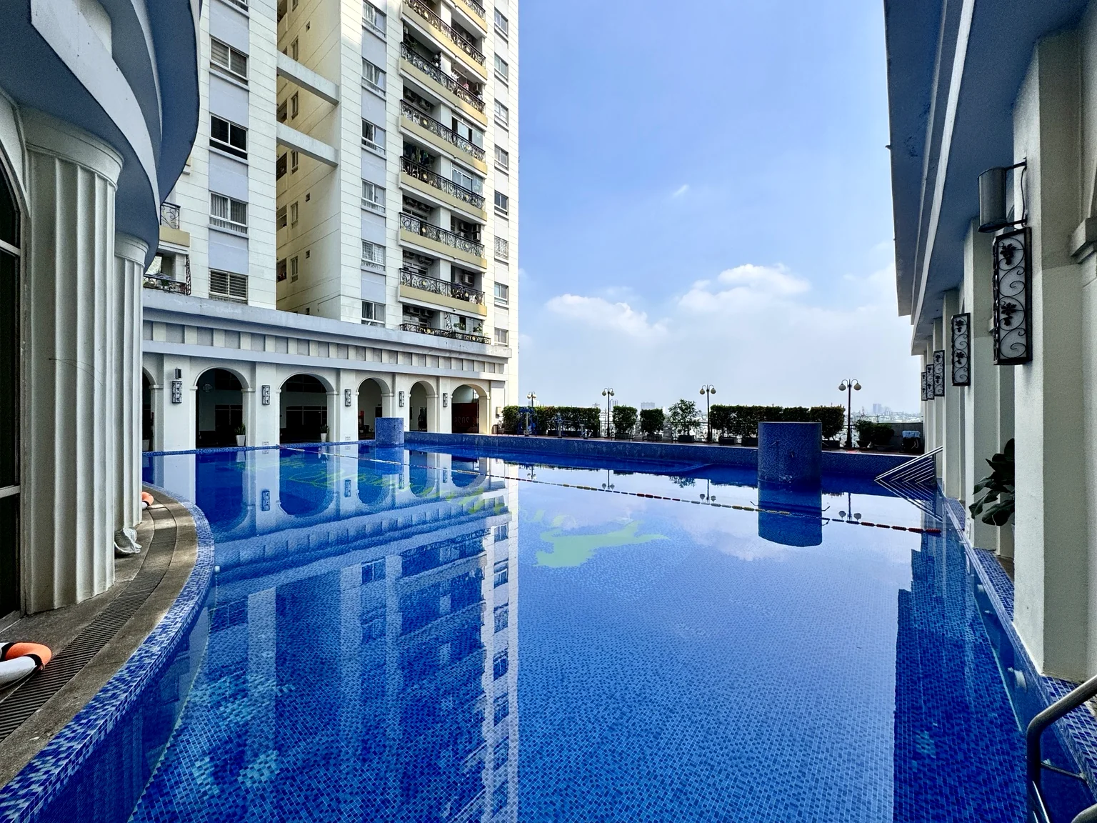 Hồ bơi xanh tại The Flemington với tòa nhà căn hộ, vòm kiến trúc và bầu trời sáng.