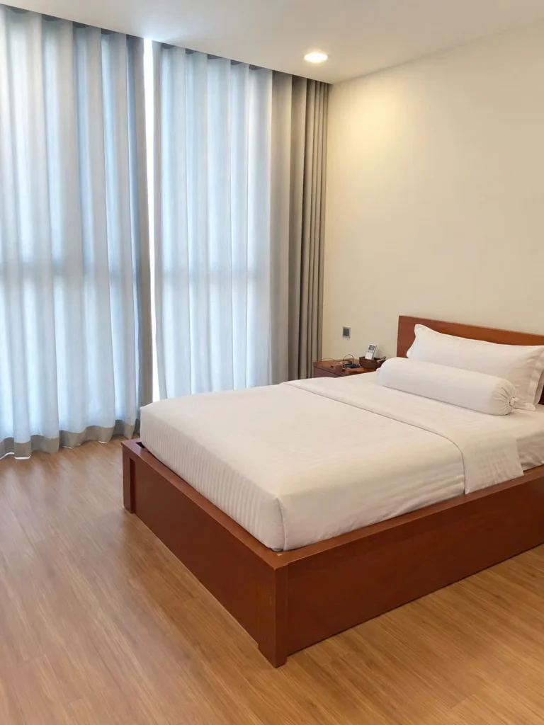 Giường gỗ phủ ga trắng cạnh cửa sổ lớn với rèm hai lớp
