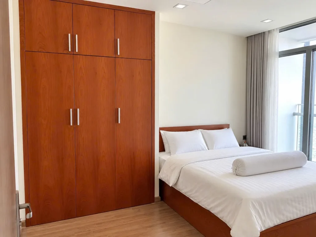 Giường ngủ cạnh cửa kính có rèm và tủ quần áo gỗ cao sát trần