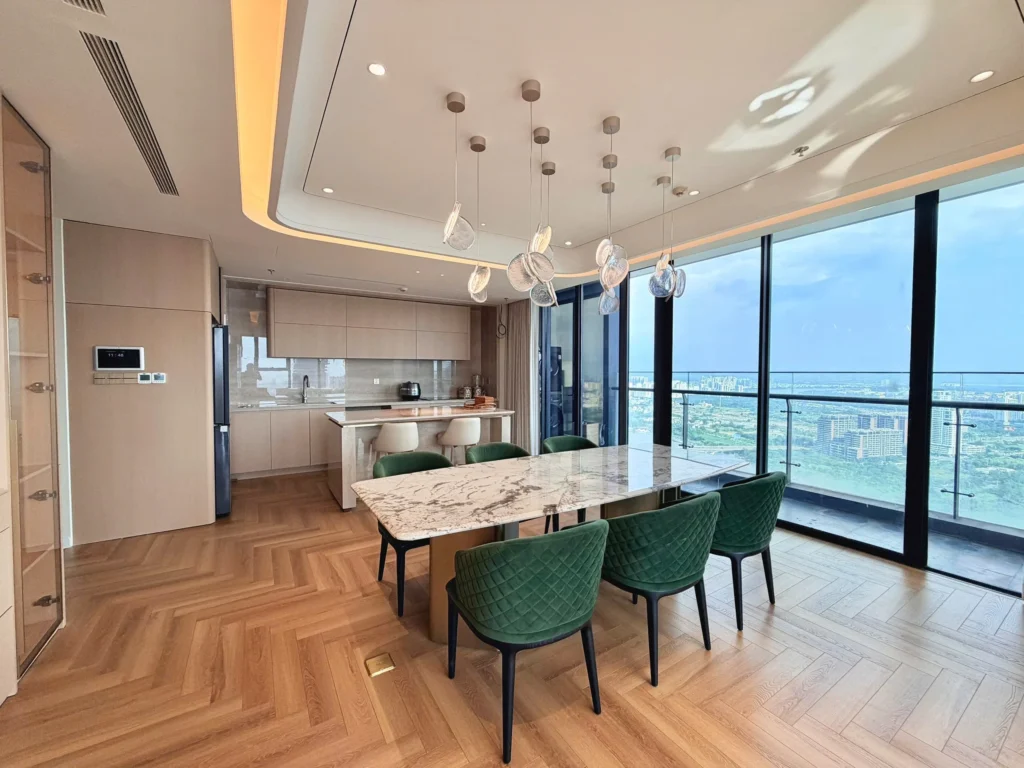 Bàn ăn mặt vân đá và ghế xanh cạnh cửa kính, phía sau là khu bếp và đảo bếp.
