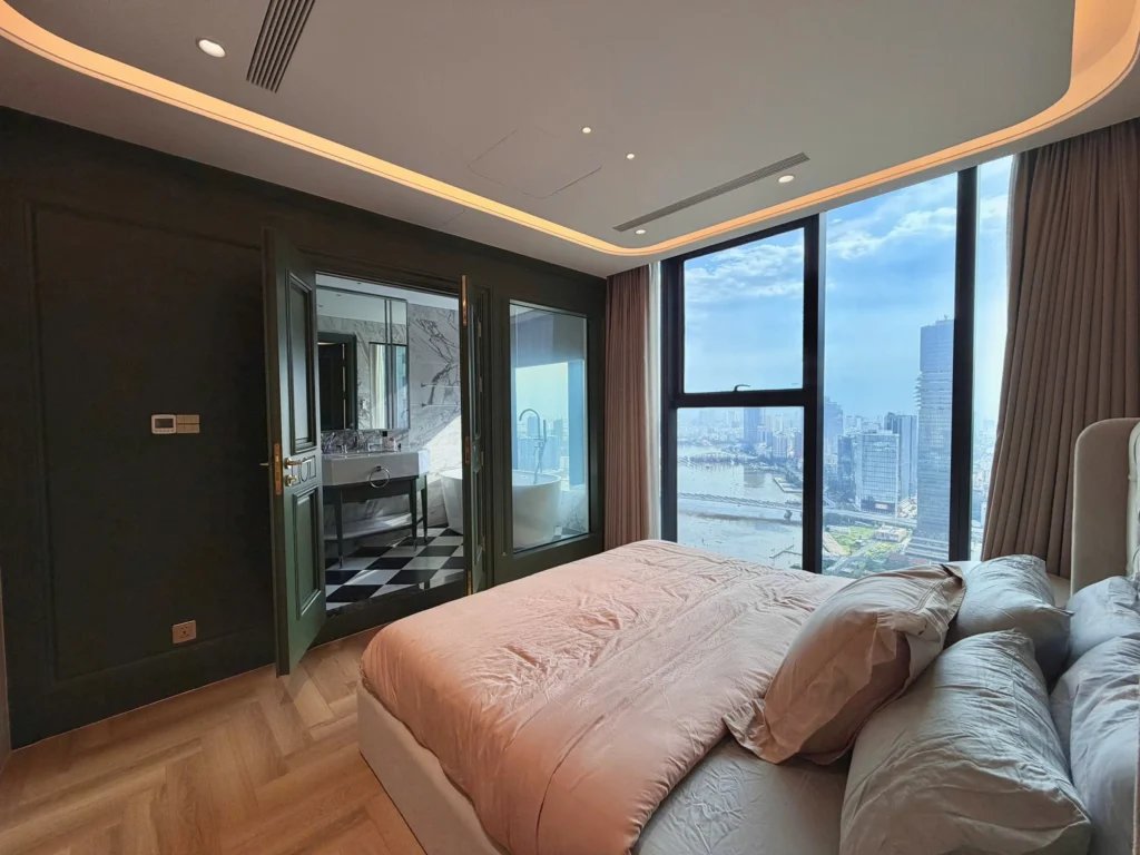 Phòng ngủ với giường lớn, hệ tủ tối màu, đèn trần và cửa kính rộng tại căn hộ Aqua 4.